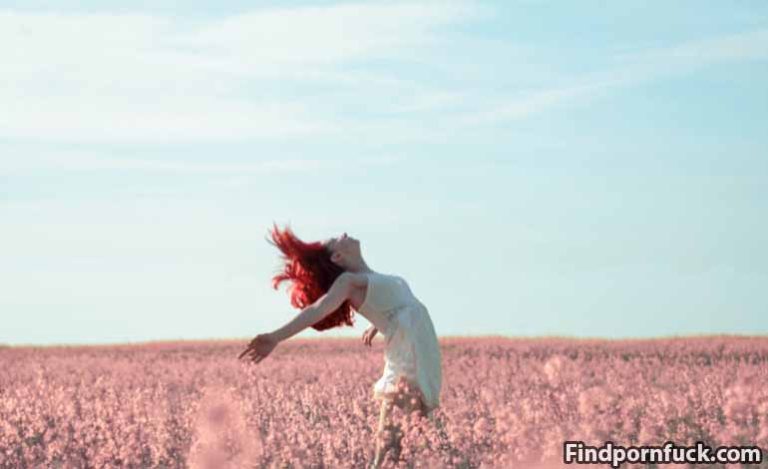 Woman with red hair in a white dress dances in a pink flower field under a blue sky, arms outstretched.